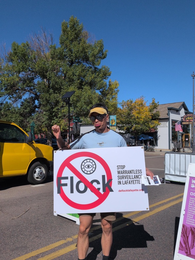 Chao wearing a sandwich-board sign reading 'Stop warrantless surveillance in Lafayette' at the farmers market